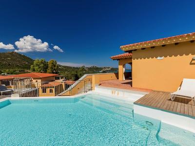 Moderner Pool mit klarem Wasser, Holzdeck und Liegestuhl vor gelbem Haus unter blauem Himmel.