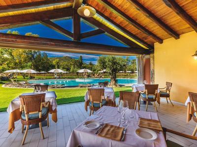 Überdachte Terrasse mit gedeckten Tischen, Blick auf grünen Garten und einen Pool mit Sonnenschirmen.
