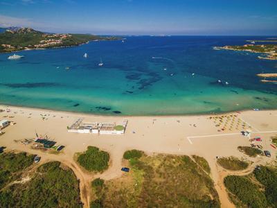 Široká písečná pláž s čistou vodou, lehátky a loděmi na horizontu.