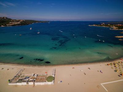 Strand mit hellem Sand, blauem Meer, Sonnenschirmen rechts und Booten auf ruhigem Wasser.