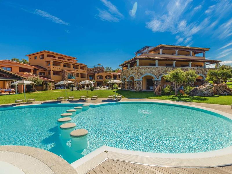 Große Villa mit mehreren Terrassen, runden Pool und grüner Rasenfläche unter blauem Himmel.