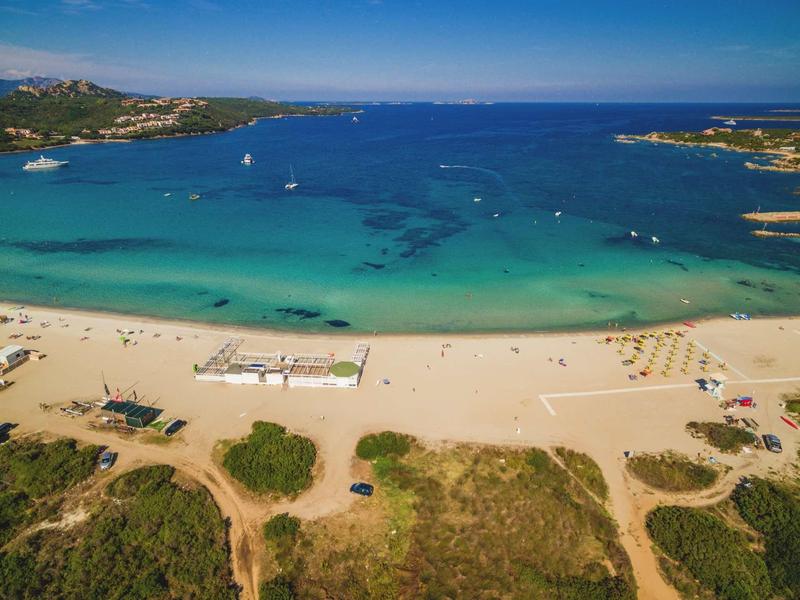 Breiter Sandstrand mit Sonnenschirmen und klarem blauem Wasser, dahinter flache grüne Hügel.