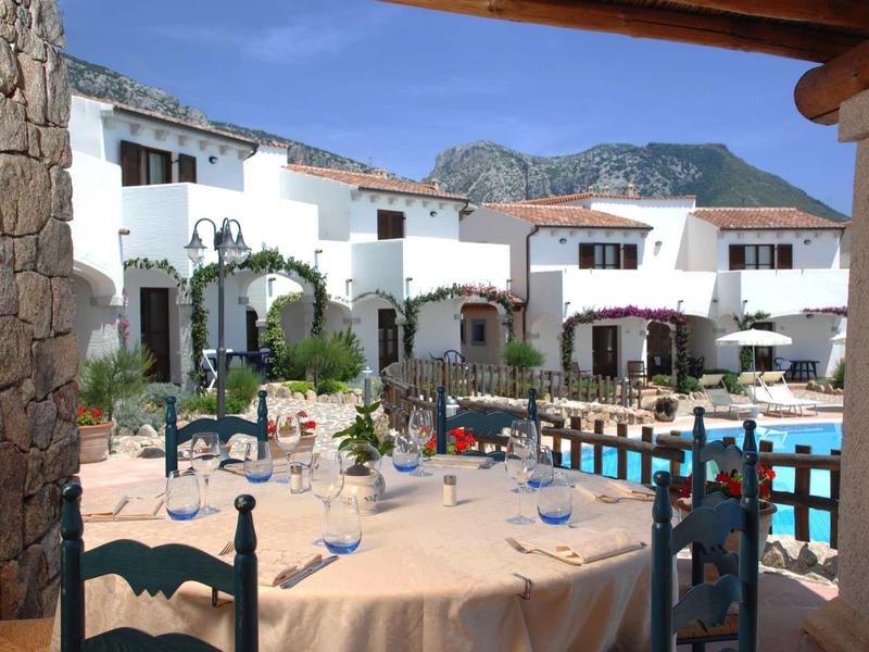 Vue sur une piscine d'hôtel et une terrasse avec des bâtiments blancs et des montagnes en arrière-plan.