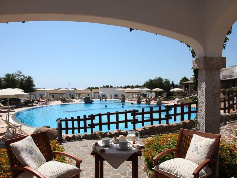 Vue depuis une terrasse couverte sur une grande piscine avec chaises longues et parasols.