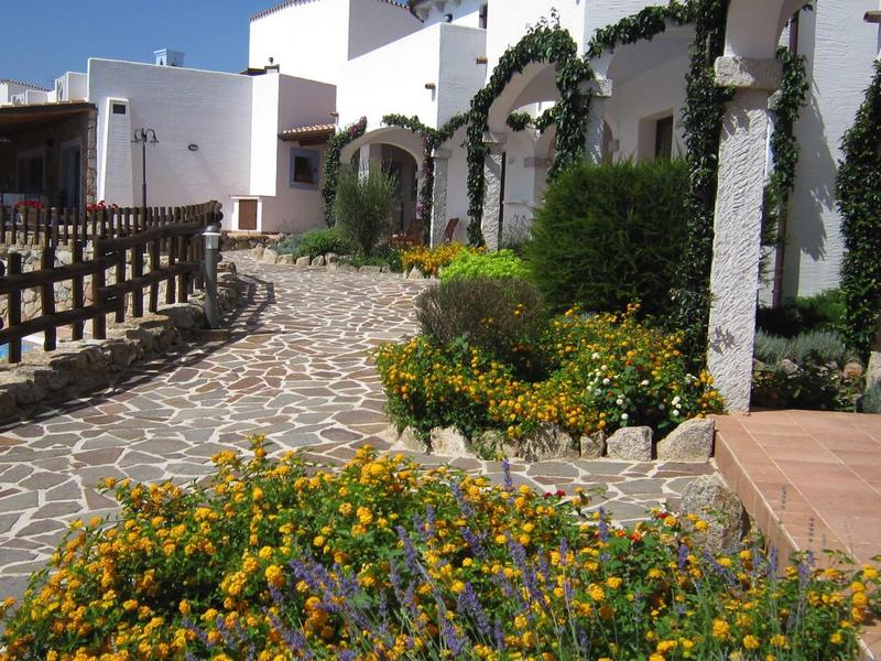 Allée pavée avec parterres de fleurs et bâtiments blanchis à la chaux sous un soleil éclatant.