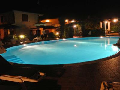 Oświetlony basen w hotelowym ogrodzie nocą z leżakami.