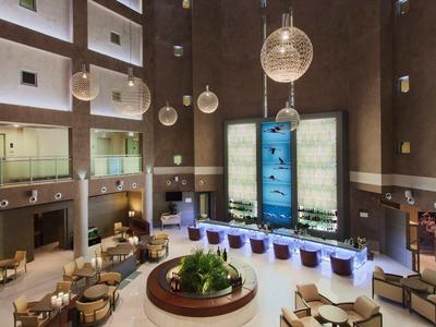 Duży hol hotelowy z wysokimi ścianami, nowoczesnymi miejscami do siedzenia i ozdobnymi lampami wiszącymi.