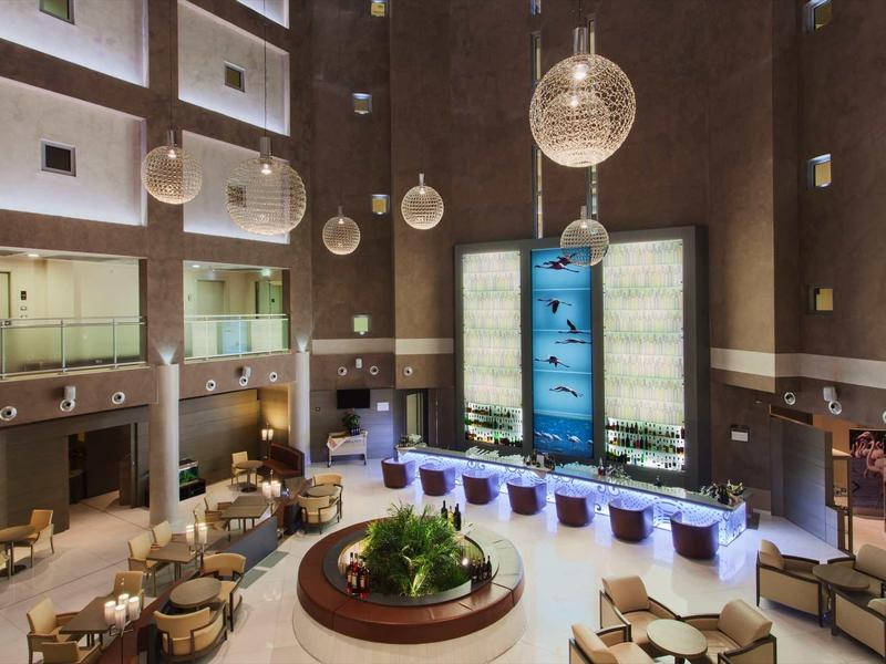 Duży hol hotelowy z wysokimi ścianami, nowoczesnymi miejscami do siedzenia i ozdobnymi lampami wiszącymi.