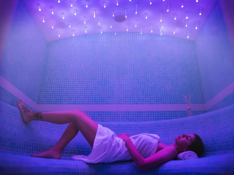 Osoba relaksująca się w łaźni parowej z niebieskimi płytkami i nastrojowym oświetleniem.