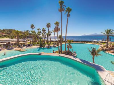 Hotelpool mit türkisfarbenem Wasser, umgeben von Palmen und Blick auf das Meer im Hintergrund.