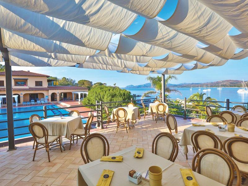 Terrasse de restaurant avec vue sur la mer et voiliers, tables avec chaises et auvents solaires.