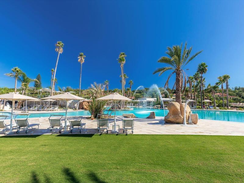 Zone de piscine d'hôtel avec parasols, chaises longues et palmiers sous un ciel bleu clair