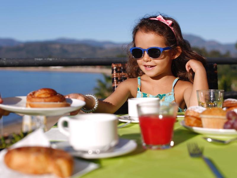 Enfant portant des lunettes de soleil profitant du petit-déjeuner sur une terrasse avec vue sur les montagnes.