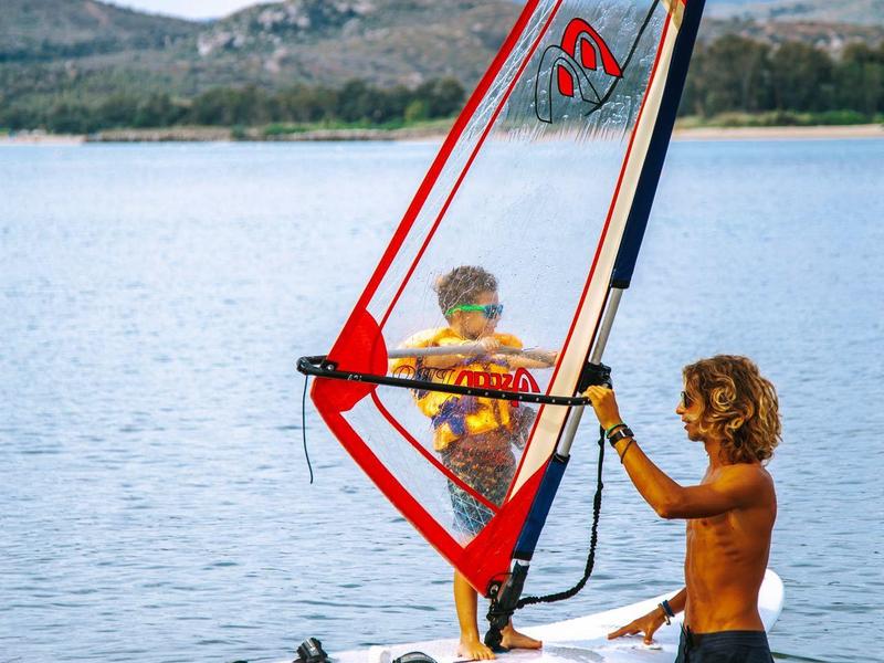 Deux enfants font de la planche à voile sur une eau calme avec des collines boisées en arrière-plan.