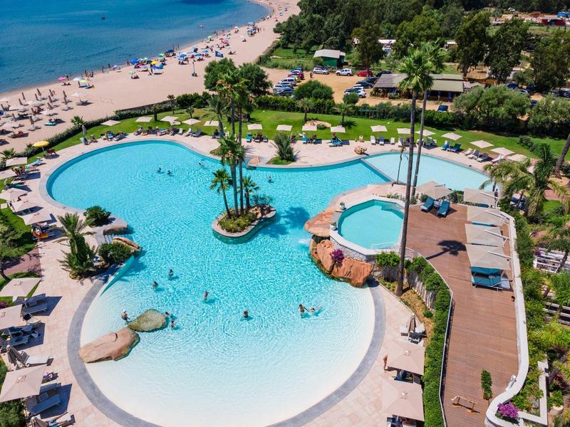 Grande piscine avec palmiers et transats à côté d'une plage de sable et de la mer bleue.