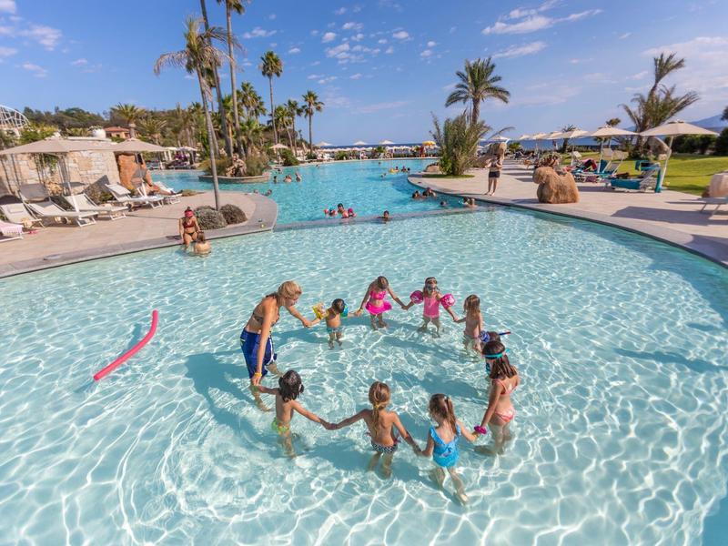 Des enfants jouent dans une piscine peu profonde d'hôtel avec des palmiers et des transats en arrière-plan.
