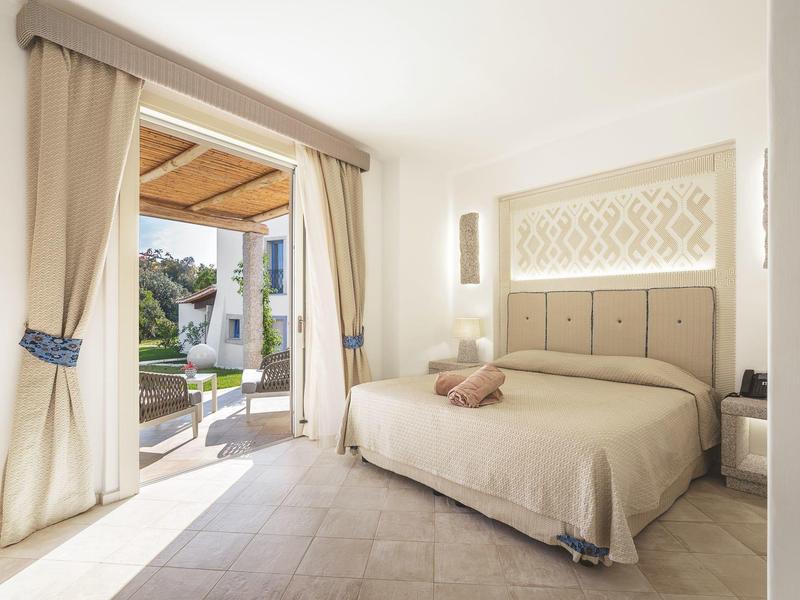 Světý hotelový pokoj s velkou postelí a otevřenými dveřmi na terasu s výhledem do zahrady.