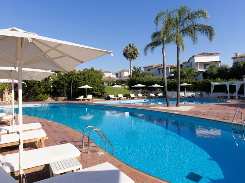 Moderní hotelový bazén s lehátky, slunečníky a palmami pod modrou oblohou.