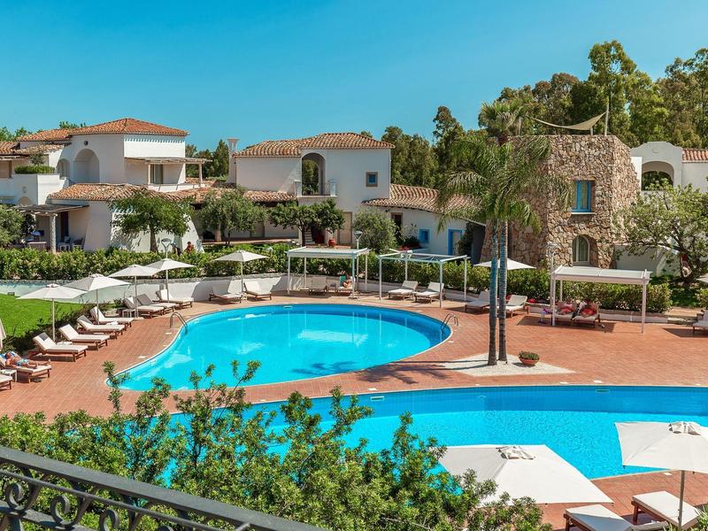 Hotelový komplex se dvěma bazény, lehátky a středomořskými budovami pod modrou oblohou.