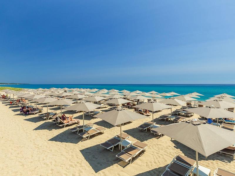 Pláž s řadami lehátek a slunečníky pod modrou oblohou u moře.