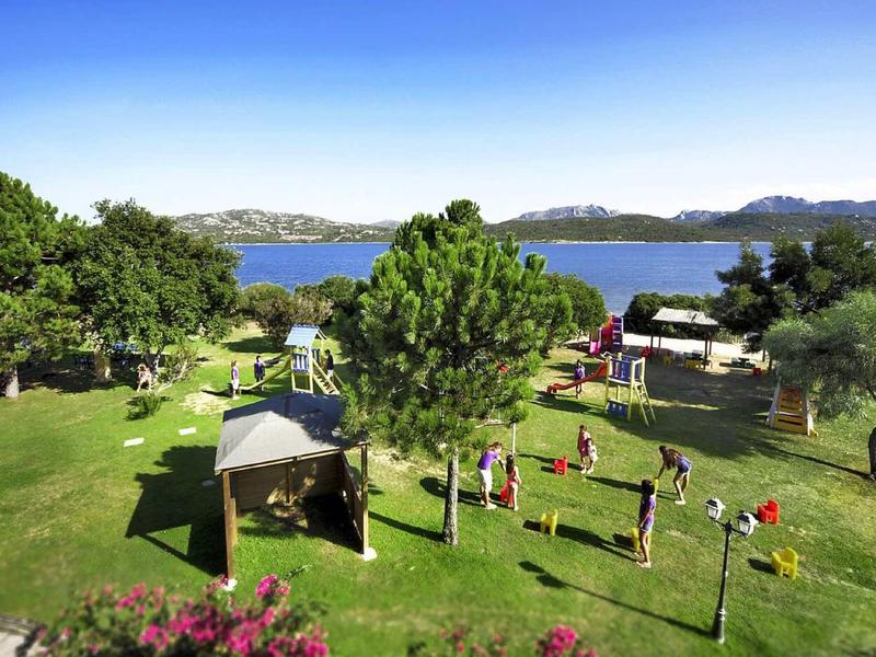 Jardin vert avec aire de jeux et vue sur lac et montagnes sous un ciel dégagé.