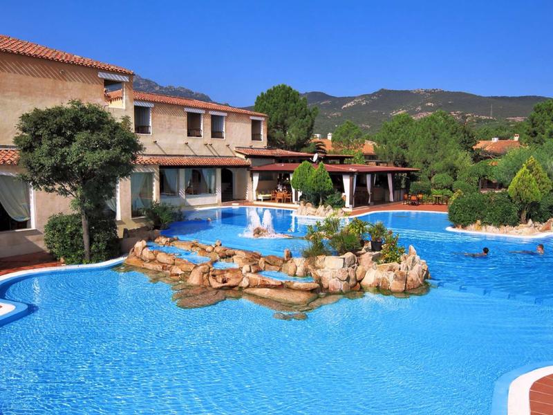 Grande piscine extérieure avec îlot rocheux devant un bâtiment hôtelier dans un paysage montagneux.
