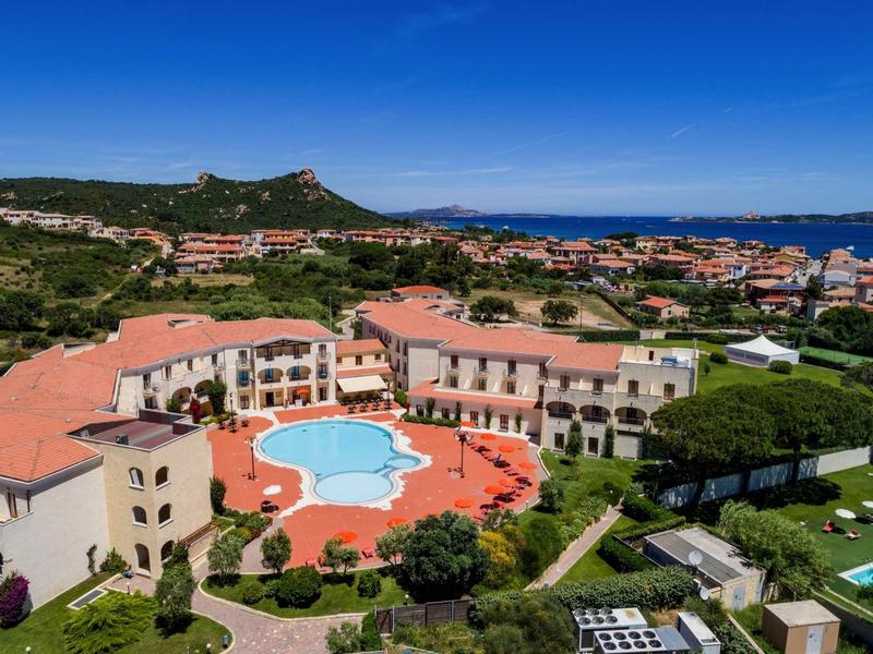 Ferienanlage mit roten Dächern, großem Pool, grünem Garten und Meer im Hintergrund unter blauem Himmel.