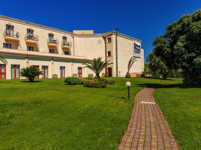 Großes gelbes Gebäude mit Balkonen, davor ein grüner Rasen und gepflasterter Weg bei blauem Himmel.