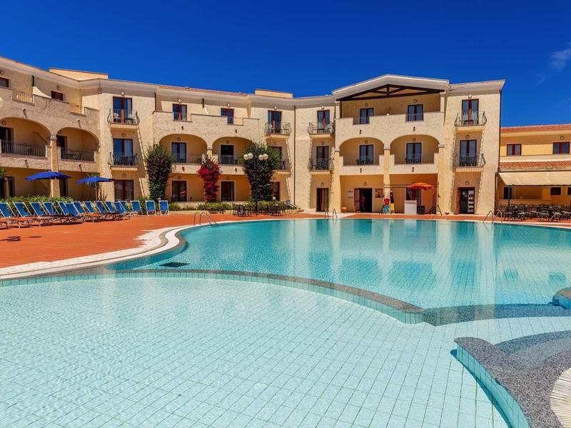 Großer, klarer Pool vor gelben Gebäuden mit Balkon und blauem Himmel im Hintergrund.