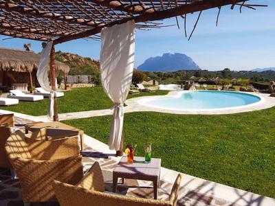Area piscina soleggiata con lettini, vista montagna e prato verde sotto un padiglione in legno.