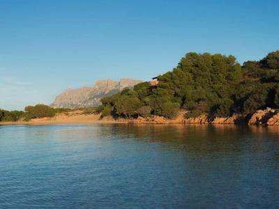 Klares blaues Wasser, bewaldete Ufer mit Felsen, im Hintergrund Berge unter blauem Himmel.
