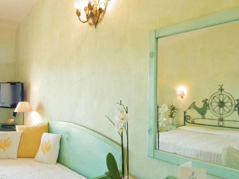 Pohodlný hotelový pokoj s pastelově zeleným nábytkem, postelí a ozdobným zrcadlem odrážejícím místnost.