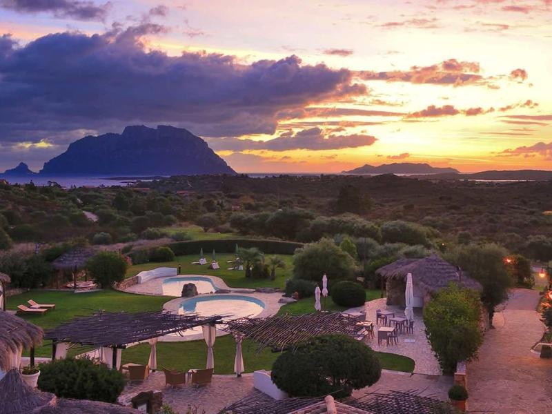 Západ slunce nad bujným resortem s bazény, palmami a vzdálenými horami pod barevnou oblohou.