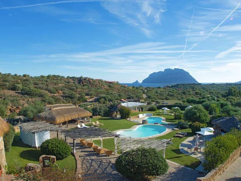 Rezort s kulatými chatkami, bazénem, zelení a horou v pozadí pod modrou oblohou.