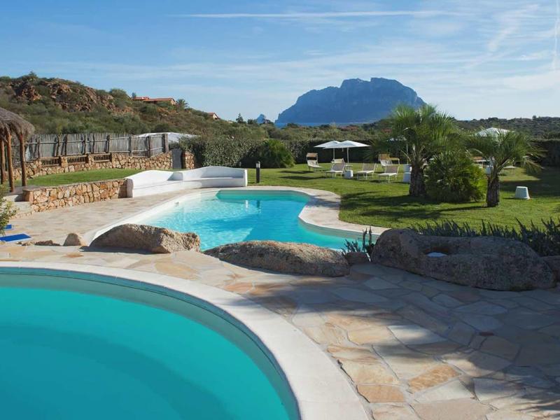 Venkovní bazén s kamennou terasou, palmami a horami v pozadí za slunečného dne.