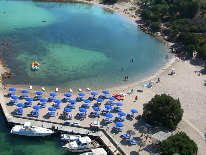 Zakřivená pláž s řadami modrých slunečníků a průzračně tyrkysovou vodou.