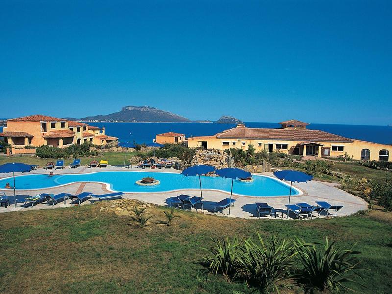 Hotelový areál s bazénem, sluncem a výhledem na moře, obklopený zelenými trávníky a rostlinami.