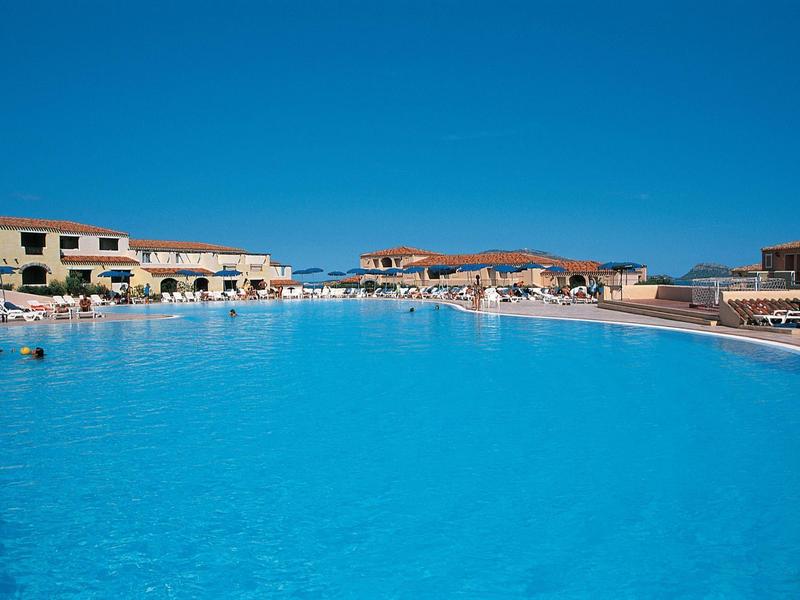 Velký čistý hotelový bazén s lehátky a jednoradými budovami pod jasnou oblohou.