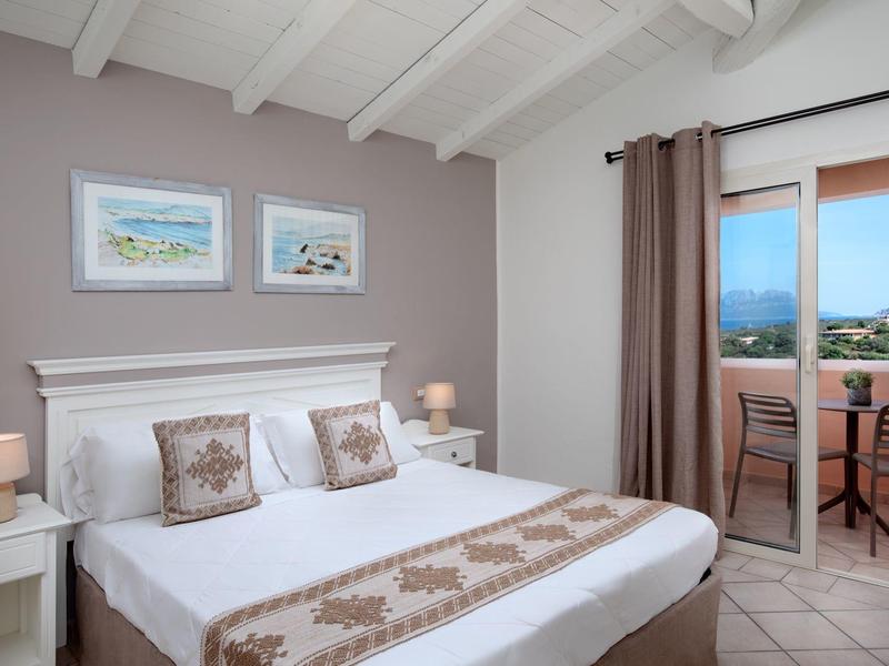 Světelná ložnice s bílou postelí, dekorativními polštáři, nočními stolky a přístupem na balkon.