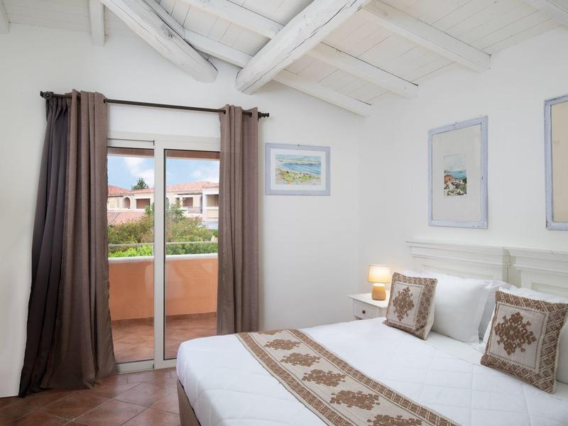 Světelná ložnice s bílými dřevěnými trámy, manželská postel a přístup na balkon s výhledem na domy.