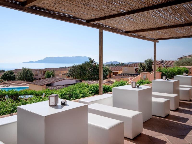 Zastřešená terasa s bílým posezením, výhled na bazén, domy a moře v pozadí.