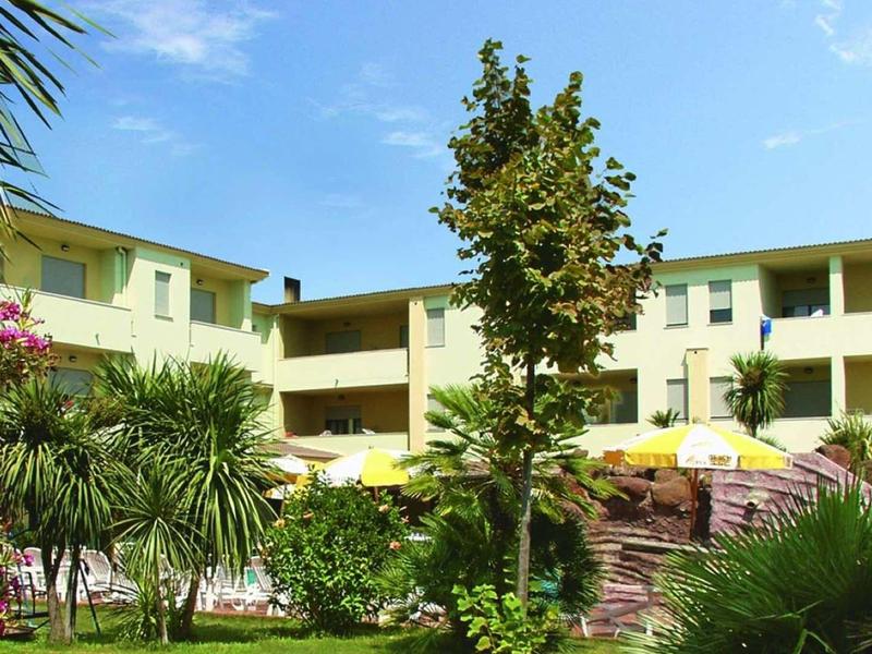 Hotel con edificio giallo, giardino con palme e fiori colorati sotto cielo blu.