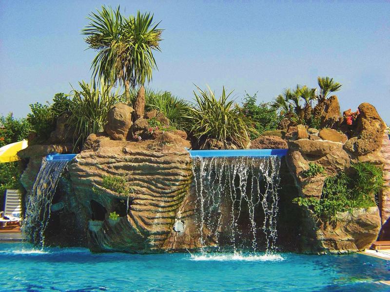 Cascata artificiale che si getta in una piscina blu chiaro con rocce e piante tropicali.