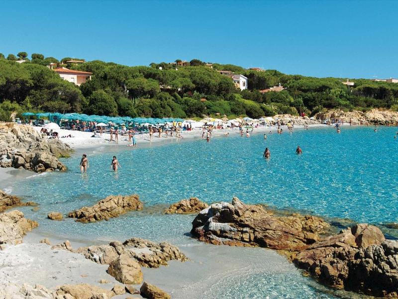 Pobřeží s skalami a čistou modrou vodou, lidé koupající se a odpočívající na pláži.