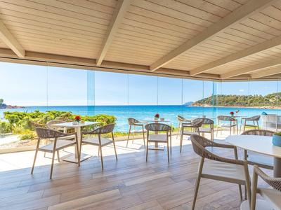 Terrazza con pavimento in legno e sedie che si affacciano sul mare blu e sulla spiaggia sotto un cielo sereno.