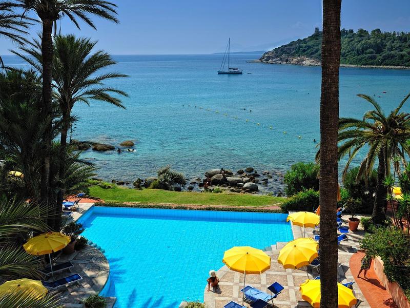 Strandhotel met zwembad, palmbomen en uitzicht op de blauwe zee met zeilboot.