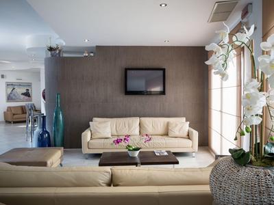 Modern eingerichtetes Wohnzimmer mit beigen Sofas, Wand-TV und großen Fenstern mit Blumen.