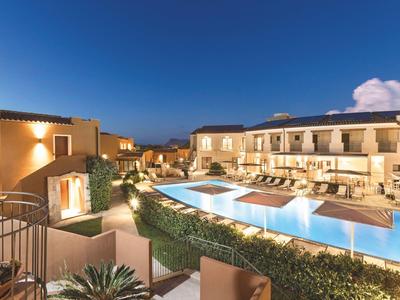 Vista serale di un elegante hotel con piscina illuminata e aree salotto all'aperto sotto un cielo sereno.