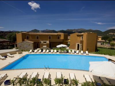 Schwimmbad mit blauen Wasser, umgeben von Liegestühlen, Sonnenschirmen und einem gelben Hotelgebäude.