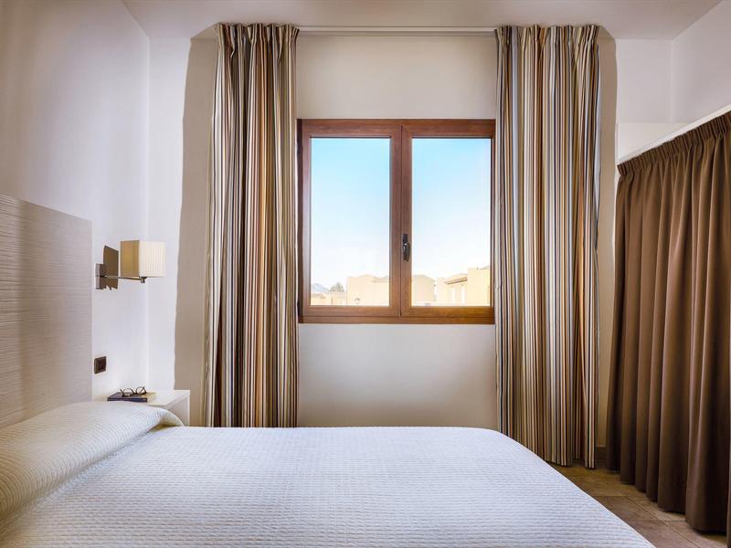 Helles Hotelzimmer mit großem Fenster, weißen Wänden und braunen Vorhängen neben einem Bett.